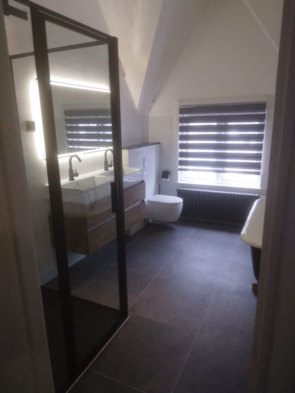 Gerenoveerde badkamer met zwart-omlijste glazen wand, houten wastafelmeubel en donkere tegelvloer
