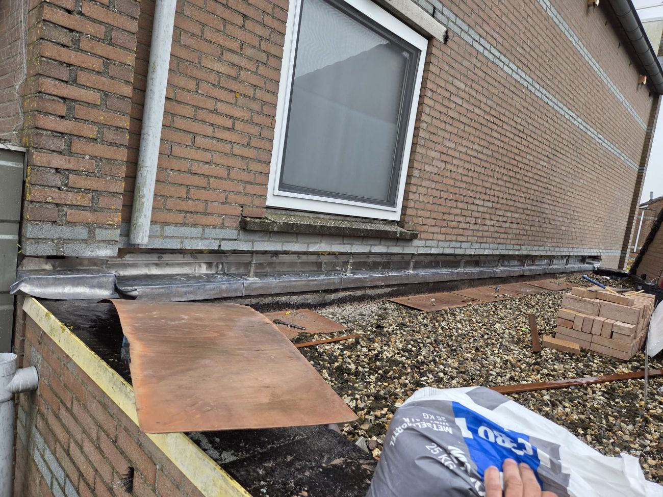 Nieuw loodwerk langs de voet van een bakstenen muur op een plat grinddak, tijdens de uitvoering.